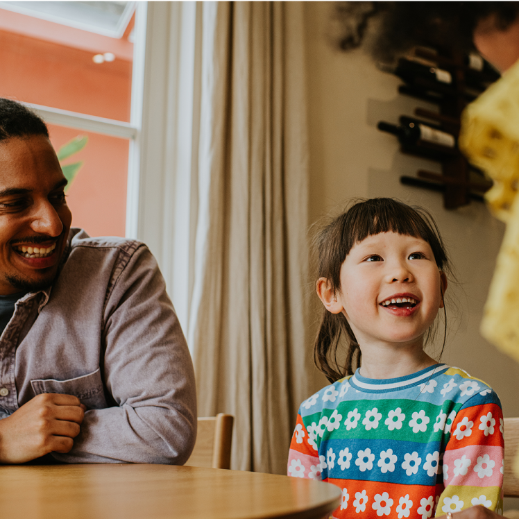 A man and two children sit at a table, radiating wellness. One child, wearing a colorful floral top, smiles warmly at the other child.