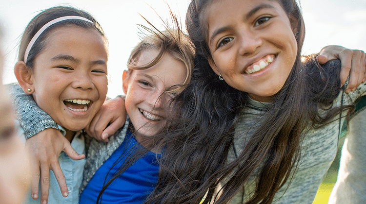 Three children stand close together outdoors, smiling and laughing at the camera on a sunny day—reminding us of the joy and connection that inspires many to become a wellness coach.
