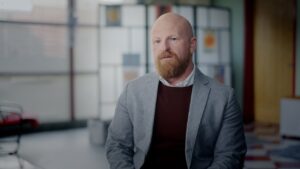 A man with a beard and bald head, dressed in a grey blazer and brown sweater, sits indoors against a blurred background of large windows and geometric decor.
