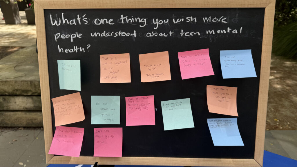 A black chalkboard with "What's one thing you wish people knew about teen mental health?" written in chalk at the top. Bright, colorful, square sticky notes with various handwriting are stuck to the board below the prompt.