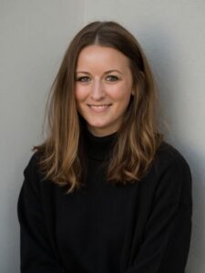 headshot of a woman with mid-length brown hair wearing a black top against a beige backdrop.